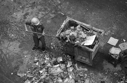 A worker wearing a safety helmet and casual clothing is standing on a paved outdoor surface next to a large, open-top skip filled with mixed waste materials, including cardboard, plastic, and other debris. The worker is holding a long-handled tool, possibly a rake or shovel, and is in the process of removing or organizing waste. In front of the skip, there is a pile of discarded rubbish, consisting of crumpled paper, plastic bags, and other small debris scattered across the ground. The surrounding environment appears to be an outdoor area within an urban setting, likely part of a property clearance in Westminster, with the surface showing signs of wear and dirt. The scene is captured in black and white, emphasizing the textures and nature of the waste and surface materials. This image relates to house clearance and rubbish removal services offered by Westminster House Clearance, demonstrating waste collection and disposal activities typical in London residential or commercial property cleaning projects.