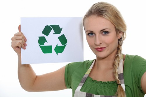 A young woman with blonde hair styled in a loose braid, wearing a green short-sleeved top and a striped apron, holds up a white sheet of paper featuring a large green recycling symbol composed of three arrows forming a triangle. She stands in front of a plain white background, with her right arm extended forward to display the recycling icon clearly, while her left arm is relaxed at her side. The image emphasizes environmental consciousness and waste recycling awareness, relevant to rubbish removal services available in Westminster or nearby areas, with a focus on sustainability and responsible disposal practices associated with Westminster House Clearance.