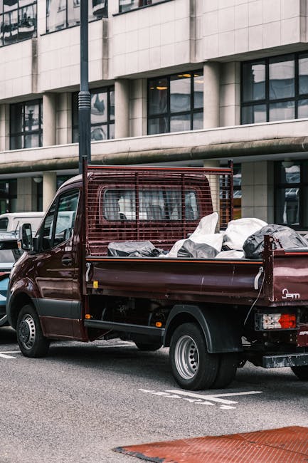A dark red flatbed pickup truck parked on an urban street in front of a modern, multi-story office building with large glass windows and a beige stone facade. The truck's bed is filled with various ty